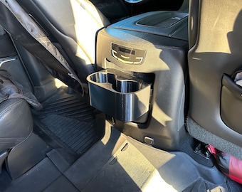 May include: Black plastic cup holder for a car. The cup holder is attached to the back of the front seat and has two cup slots.