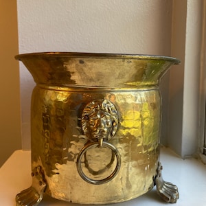 May include: A hammered brass planter featuring a wide, flared rim and a cylindrical body. The exterior is adorned with a prominent lion head motif holding a circular ring handle. The vessel is supported by three decorative claw-style feet.