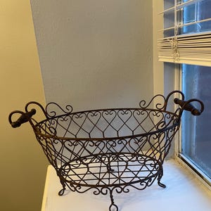 May include: An oval-shaped wire basket with a dark brown, rusted finish. The basket features a decorative lattice pattern along the sides, two curved handles on opposite ends, and four small, curled metal feet supporting the base structure.