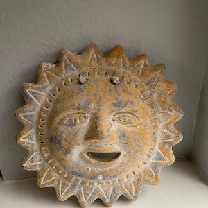 May include: A circular sun-shaped wall plaque made of textured, weathered clay. The piece features a central face with a smiling mouth, almond-shaped eyes, and a radiating sunburst border with triangular points in shades of ochre and blue.