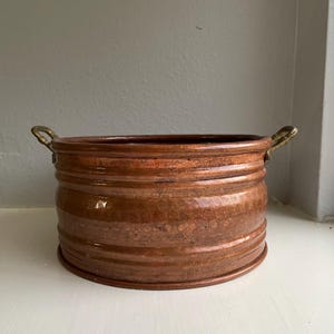 May include: A round, hammered copper basin featuring a wide, cylindrical body with horizontal ribbed detailing and two small, curved brass handles attached to opposite sides. The metal surface displays a warm, metallic brown patina throughout.