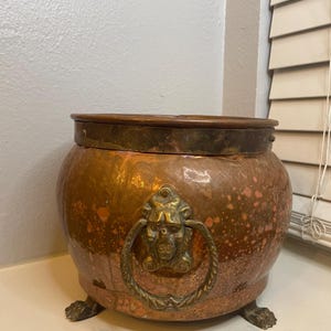 May include: A copper-colored, round pot with a bronze rim and a decorative bronze handle featuring a face. The pot has three claw feet and is likely used for decorative purposes.