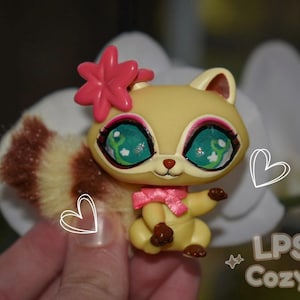 May include: A yellow squirrel figurine with large, detailed teal eyes and a pink bow. It has a pink flower on its head and a fluffy, striped tail. The text "LPS Cozy" is visible in the lower right corner. The figurine is a collectible toy.