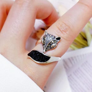 May include: A silver fox ring featuring a black glitter tail and a detailed fox head. The ring is worn on a finger, highlighting the intricate design. The ring's design is a unique piece of jewellery.