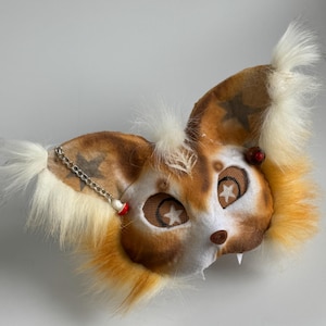 May include: A handmade mask in brown, white, and orange, resembling a fox. The mask has star-shaped eye details, a chain, and small fangs. The mask is designed for costume or cosplay use. The mask is made of faux fur and fabric.