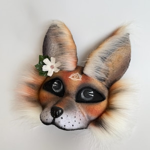 May include: A fox-themed mask featuring faux fur in shades of orange, white, and black. The mask has large black mesh eyes, a black nose, and a white flower accessory on the left ear. A small white triangle symbol is painted on the forehead.