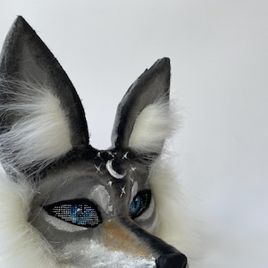 May include: A gray and white wolf mask with blue eyes and a crescent moon painted on the forehead. The mask has white fur around the edges.