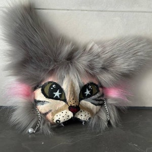 Puede incluir: Una cabeza de gato de peluche gris con orejas rosas y una cara pintada. El gato tiene ojos verdes con estrellas blancas, una nariz roja y una boca negra. El gato lleva una cadena plateada con una perla colgando.