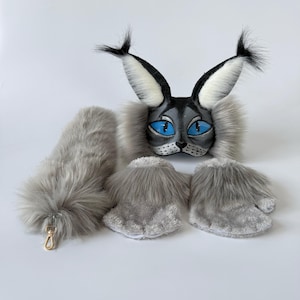 May include: A lynx-inspired costume set featuring a grey and black mask with large pointed ears, bright blue eyes, and long white tufts. The set includes a long, fluffy grey tail with a gold-toned metal clasp and two grey faux fur paw-style mittens.