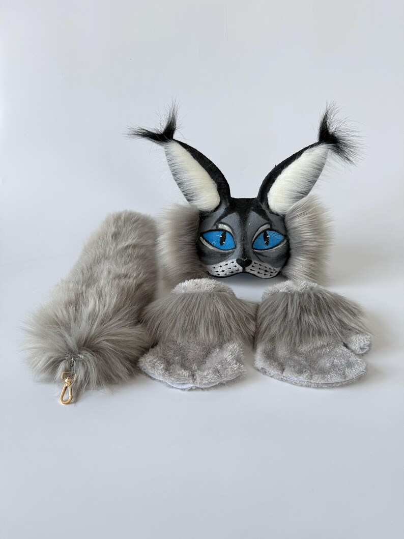 May include: A lynx-inspired costume set featuring a grey and black mask with large pointed ears, bright blue eyes, and long white tufts. The set includes a long, fluffy grey tail with a gold-toned metal clasp and two grey faux fur paw-style mittens.