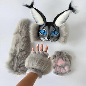 May include: A set consisting of a grey faux fur cat mask with bright blue eyes and black-tipped ears, a long grey faux fur tail, a pair of grey faux fur fingerless gloves, and a small grey faux fur pouch featuring a pink paw print design.