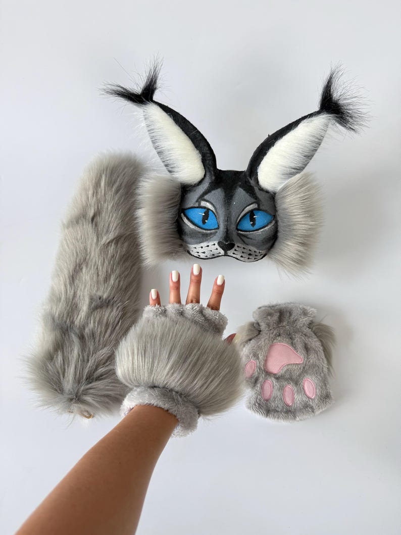 May include: A set consisting of a grey faux fur cat mask with bright blue eyes and black-tipped ears, a long grey faux fur tail, a pair of grey faux fur fingerless gloves, and a small grey faux fur pouch featuring a pink paw print design.
