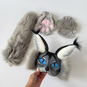 May include: A gray faux fur cat costume set including a mask with large blue eyes and tall tufted ears, a long fluffy tail, and two fingerless gloves featuring pink paw pads. The mask has black and white fur accents and a detailed feline face.