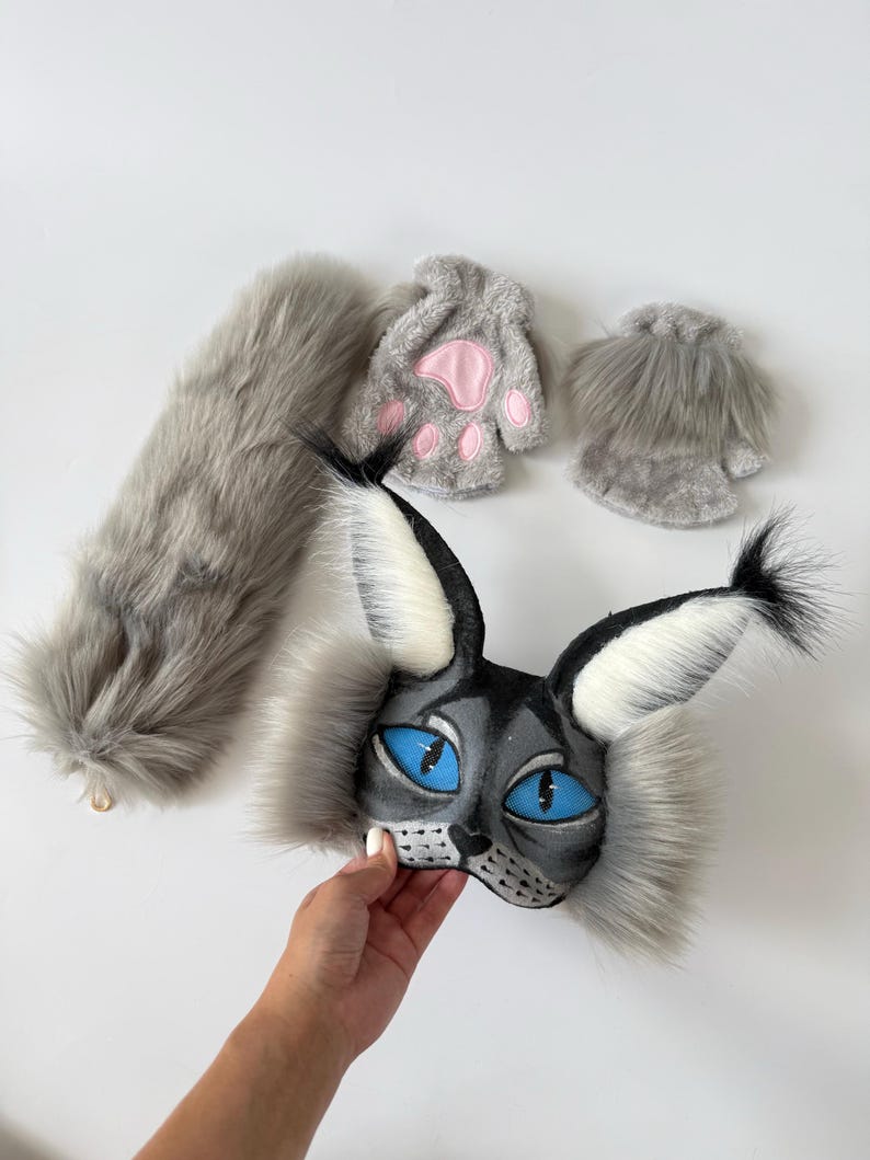 May include: A gray faux fur cat costume set including a mask with large blue eyes and tall tufted ears, a long fluffy tail, and two fingerless gloves featuring pink paw pads. The mask has black and white fur accents and a detailed feline face.
