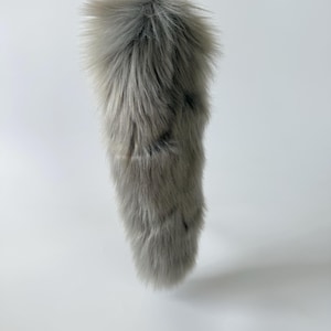May include: A fluffy, light gray faux fur tail keychain featuring long, soft synthetic fibers with subtle darker gray streaks throughout. The accessory is attached to a gold-toned metal lobster clasp, allowing it to be easily clipped onto bags or keys.