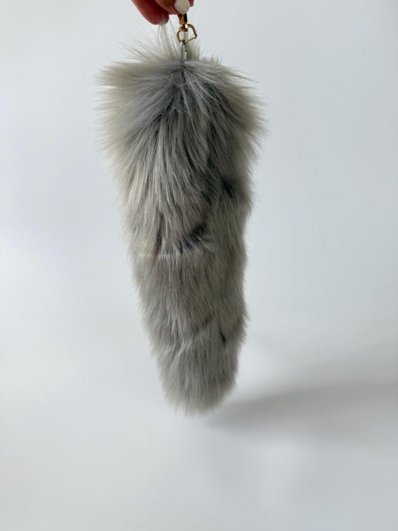 May include: A fluffy, light gray faux fur tail keychain featuring long, soft synthetic fibers with subtle darker gray streaks throughout. The accessory is attached to a gold-toned metal lobster clasp, allowing it to be easily clipped onto bags or keys.