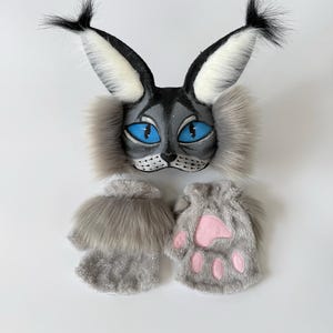 May include: A gray and white cat-themed mask featuring tall tufted ears, bright blue eyes, and long faux fur side whiskers, paired with two gray fingerless gloves that have soft faux fur cuffs and pink paw pad details on the palms.
