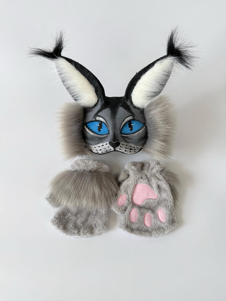 May include: A gray and white cat-themed mask featuring tall tufted ears, bright blue eyes, and long faux fur side whiskers, paired with two gray fingerless gloves that have soft faux fur cuffs and pink paw pad details on the palms.