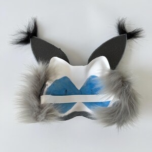 May include: A white mask featuring two tall, pointed dark gray ears with black tufts at the tips. The mask has fluffy gray faux fur accents on the sides and a white elastic strap across the back, with blue mesh fabric covering the eye openings.