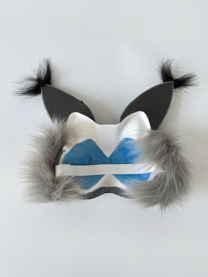 May include: A white mask featuring two tall, pointed dark gray ears with black tufts at the tips. The mask has fluffy gray faux fur accents on the sides and a white elastic strap across the back, with blue mesh fabric covering the eye openings.