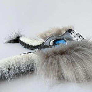 May include: A handcrafted mask featuring long, soft grey and white faux fur. The mask has a sculpted snout with black painted details, bright blue eye accents, and a pointed ear structure. The design incorporates a long, furry tail-like extension.