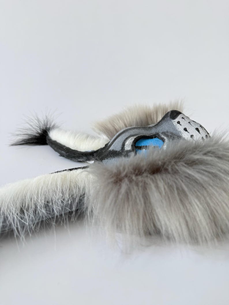 May include: A handcrafted mask featuring long, soft grey and white faux fur. The mask has a sculpted snout with black painted details, bright blue eye accents, and a pointed ear structure. The design incorporates a long, furry tail-like extension.