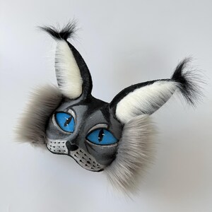 May include: A cat-themed mask featuring grey and black painted details, vibrant blue mesh eyes, and long, fluffy grey and white faux fur accents on the cheeks and tall, pointed ears with black tufted tips at the top of each ear.