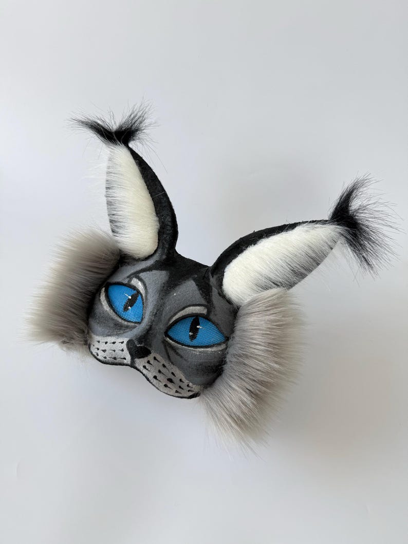 May include: A cat-themed mask featuring grey and black painted details, vibrant blue mesh eyes, and long, fluffy grey and white faux fur accents on the cheeks and tall, pointed ears with black tufted tips at the top of each ear.
