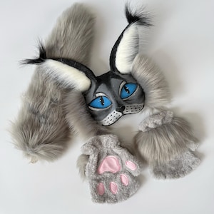 May include: A gray and white faux fur set consisting of a cat-like mask with pointed ears, bright blue eyes, and black tufts, a long fluffy tail, and two fingerless paw gloves featuring pink padded details on the palms and gray fur.