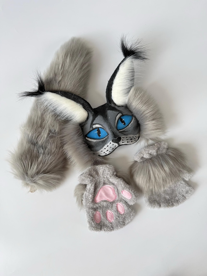 May include: A gray and white faux fur set consisting of a cat-like mask with pointed ears, bright blue eyes, and black tufts, a long fluffy tail, and two fingerless paw gloves featuring pink padded details on the palms and gray fur.