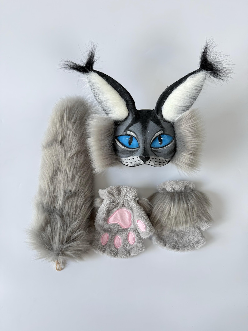 May include: A gray and black faux fur animal mask with large pointed ears, bright blue eyes, and whiskers. The set includes a long, fluffy gray tail and two gray fingerless gloves featuring pink paw pads on the palms and furry cuffs.