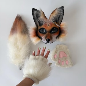 May include: A fox-themed costume set featuring a mask with orange, black, and white faux fur, a silver chain detail, and a painted eye symbol on the forehead. The set includes a long, fluffy white and orange tail and two white faux fur paw gloves.