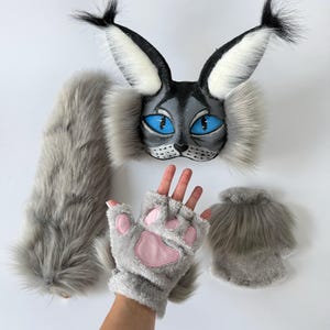 May include: A gray and black faux fur cat mask with large pointed ears, tufted tips, and bright blue eyes. The set includes a long gray faux fur tail and a pair of fingerless gray gloves featuring soft pink paw pads on the palm side.