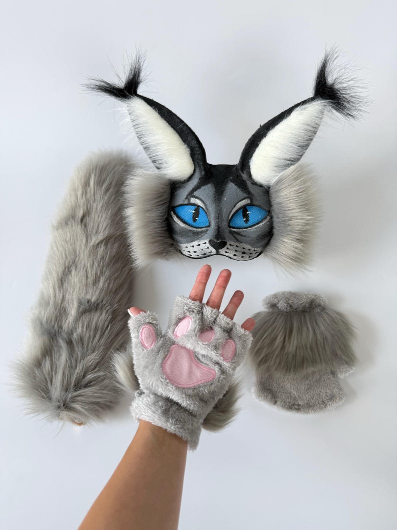 May include: A gray and black faux fur cat mask with large pointed ears, tufted tips, and bright blue eyes. The set includes a long gray faux fur tail and a pair of fingerless gray gloves featuring soft pink paw pads on the palm side.