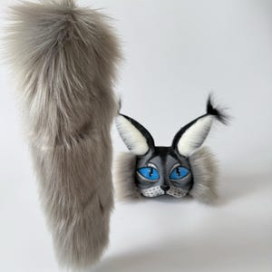 May include: A long, fluffy grey faux fur tail with a gold-toned metal lobster clasp, paired with a matching grey and black cat mask featuring pointed ears with black tufts, white inner ear fur, and bright blue painted eyes.
