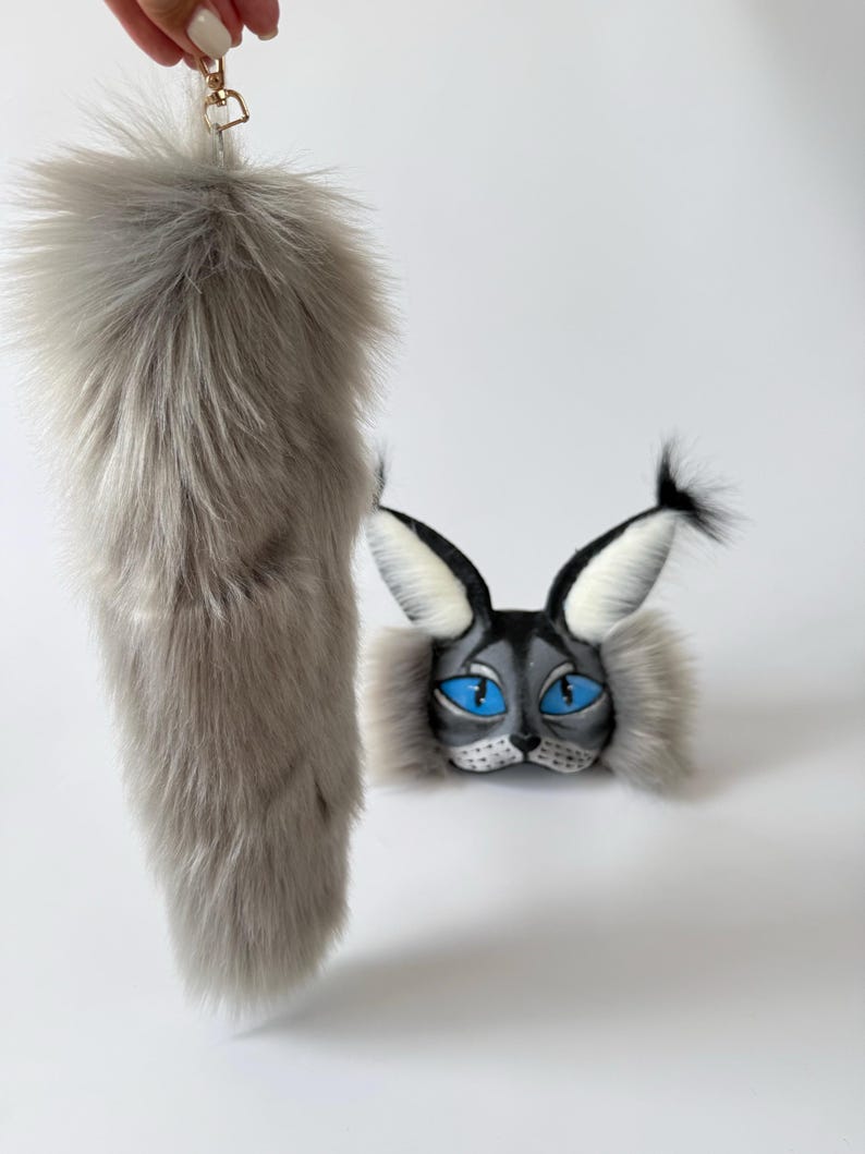 May include: A long, fluffy grey faux fur tail with a gold-toned metal lobster clasp, paired with a matching grey and black cat mask featuring pointed ears with black tufts, white inner ear fur, and bright blue painted eyes.