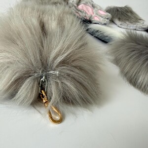 May include: A set of gray faux fur accessories including a long tail with a gold-toned metal lobster clasp, and a pair of fingerless gloves featuring gray fur and pink paw pad accents. The items are made of soft, long-pile synthetic gray fur.