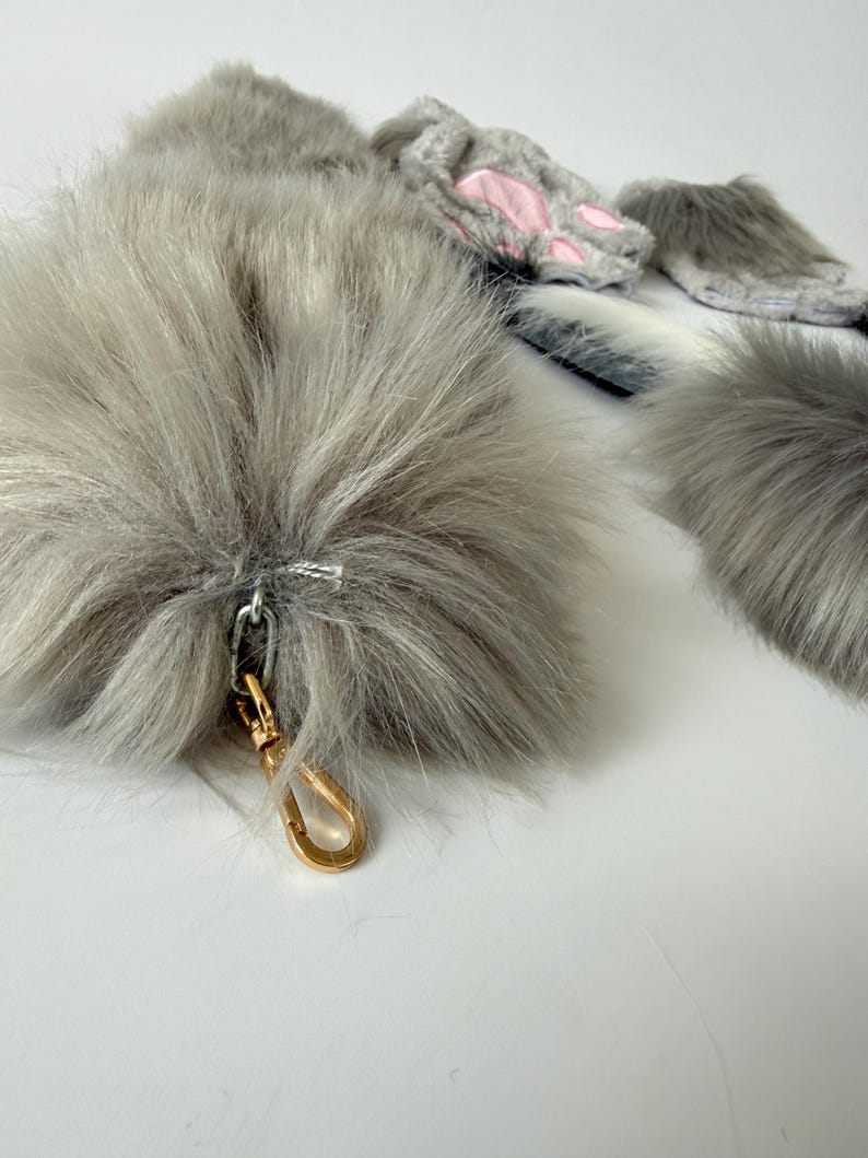 May include: A set of gray faux fur accessories including a long tail with a gold-toned metal lobster clasp, and a pair of fingerless gloves featuring gray fur and pink paw pad accents. The items are made of soft, long-pile synthetic gray fur.
