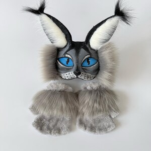 May include: A lynx-inspired mask featuring pointed ears with black tufts, bright blue eyes, and grey faux fur accents, paired with a set of matching grey faux fur fingerless gloves. The mask has a grey face with black whiskers and nose details.