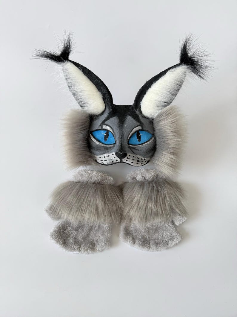 May include: A lynx-inspired mask featuring pointed ears with black tufts, bright blue eyes, and grey faux fur accents, paired with a set of matching grey faux fur fingerless gloves. The mask has a grey face with black whiskers and nose details.