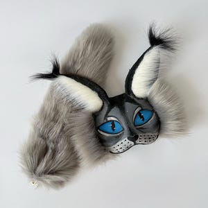 May include: A lynx-inspired mask and tail set featuring long, soft grey faux fur. The mask has a black face with bright blue almond-shaped eyes, white inner ear tufts, and black ear tips. The long, fluffy grey tail complements the mask.