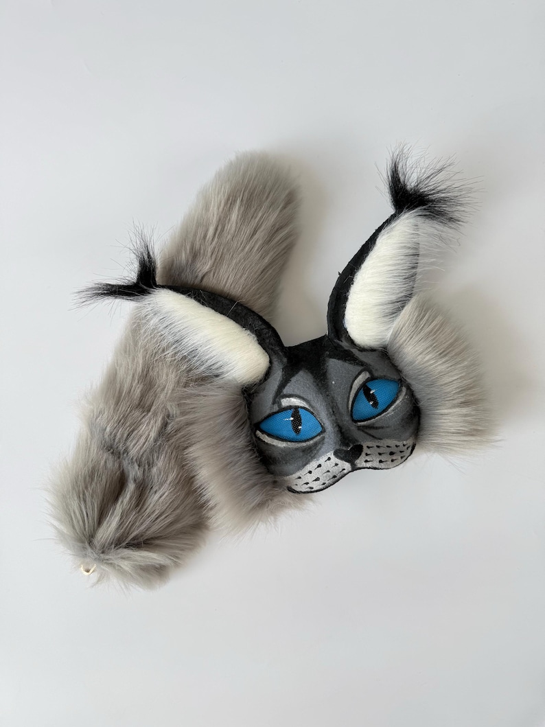 May include: A lynx-inspired mask and tail set featuring long, soft grey faux fur. The mask has a black face with bright blue almond-shaped eyes, white inner ear tufts, and black ear tips. The long, fluffy grey tail complements the mask.