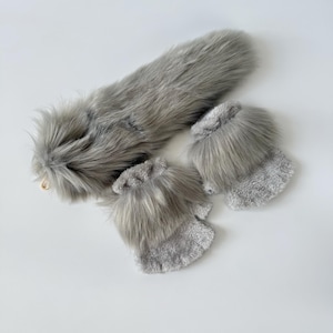 May include: A set consisting of a long, grey faux fur tail with a gold-toned metal clip and a pair of fingerless gloves. The gloves are crafted from a soft, textured grey fabric and feature a thick band of matching grey faux fur at the cuffs.
