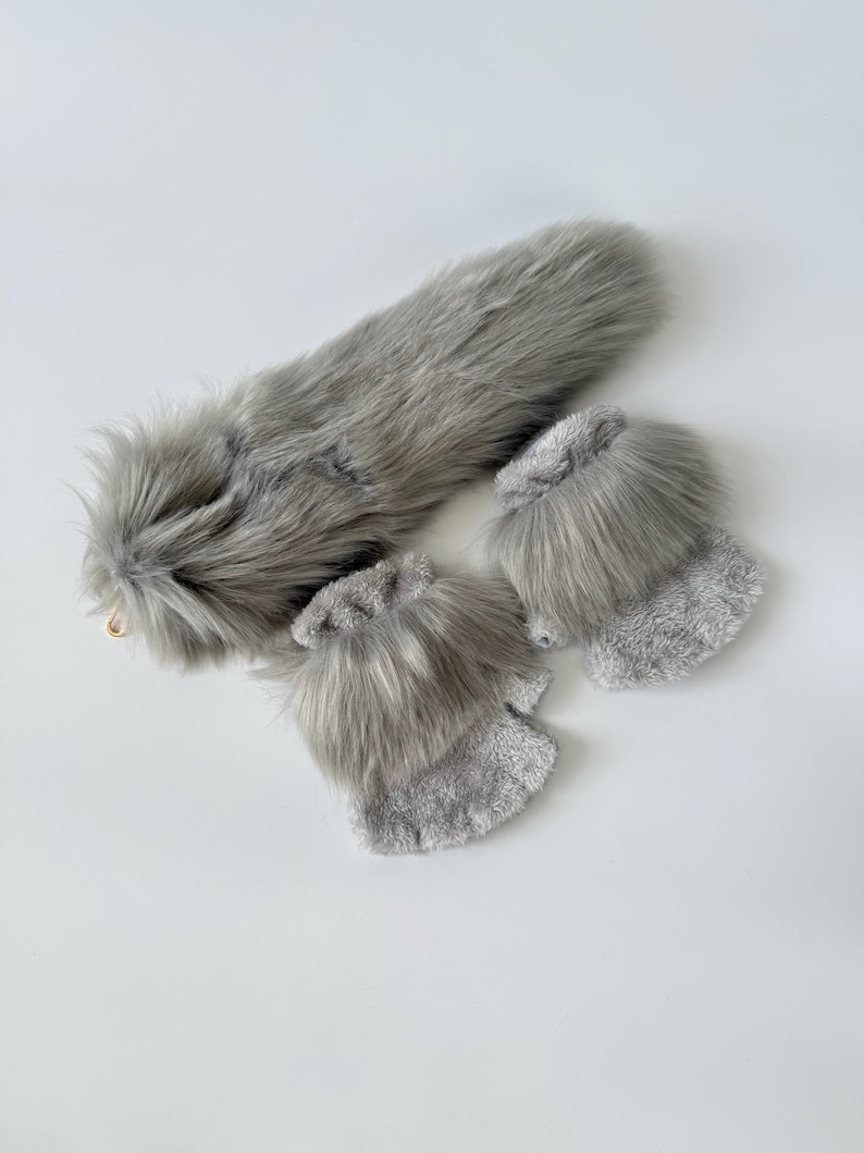 May include: A set consisting of a long, grey faux fur tail with a gold-toned metal clip and a pair of fingerless gloves. The gloves are crafted from a soft, textured grey fabric and feature a thick band of matching grey faux fur at the cuffs.