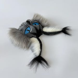 May include: A sculpted mask featuring a dark gray face with bright blue mesh eyes, accented by long, fluffy gray and white faux fur tufts extending from the sides and bottom, creating a textured, layered appearance with a pointed, animal-like shape.
