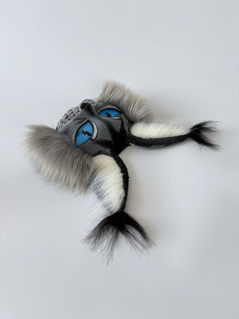 May include: A sculpted mask featuring a dark gray face with bright blue mesh eyes, accented by long, fluffy gray and white faux fur tufts extending from the sides and bottom, creating a textured, layered appearance with a pointed, animal-like shape.