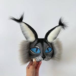 May include: A cat mask featuring a grey and black face with bright blue eyes, a black nose, and whiskers. The mask includes tall, pointed ears with black tufts at the tips and long, fluffy grey fur accents on the sides of the face.