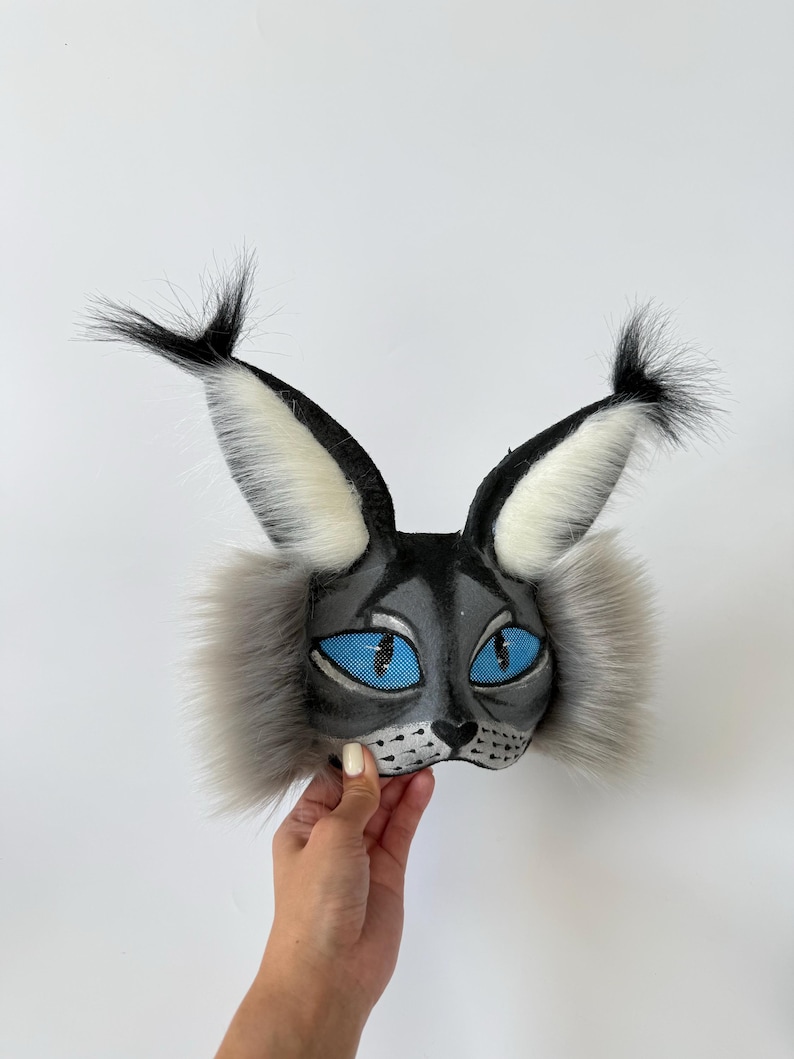 May include: A cat mask featuring a grey and black face with bright blue eyes, a black nose, and whiskers. The mask includes tall, pointed ears with black tufts at the tips and long, fluffy grey fur accents on the sides of the face.