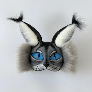 May include: A cat-themed mask featuring a grey painted face with bright blue eyes, black whiskers, and long, pointed ears topped with black tufts of faux fur. The sides of the mask are adorned with thick, grey faux fur cheek accents.