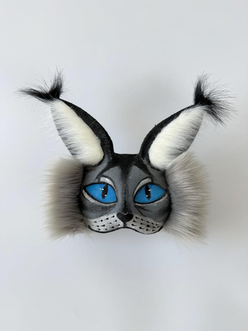 May include: A cat-themed mask featuring a grey painted face with bright blue eyes, black whiskers, and long, pointed ears topped with black tufts of faux fur. The sides of the mask are adorned with thick, grey faux fur cheek accents.